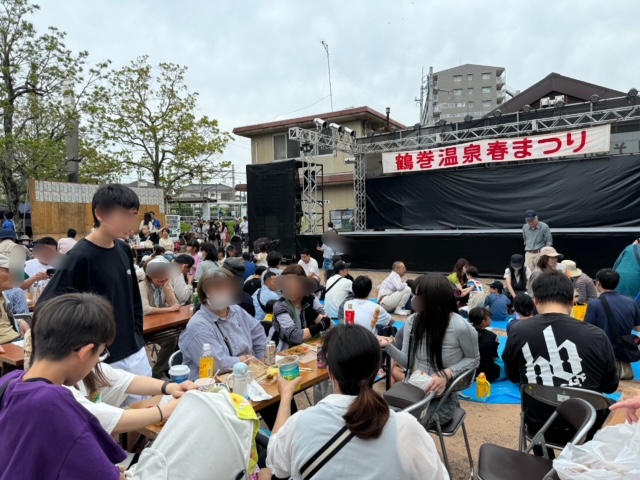 手前のテーブル席や地面に敷かれた青いシートの上で人々が食事や交流を楽しんでおり、奥には「鶴巻温泉春まつり」と書かれた横断幕が掲げられた特設ステージが設置されている会場の写真