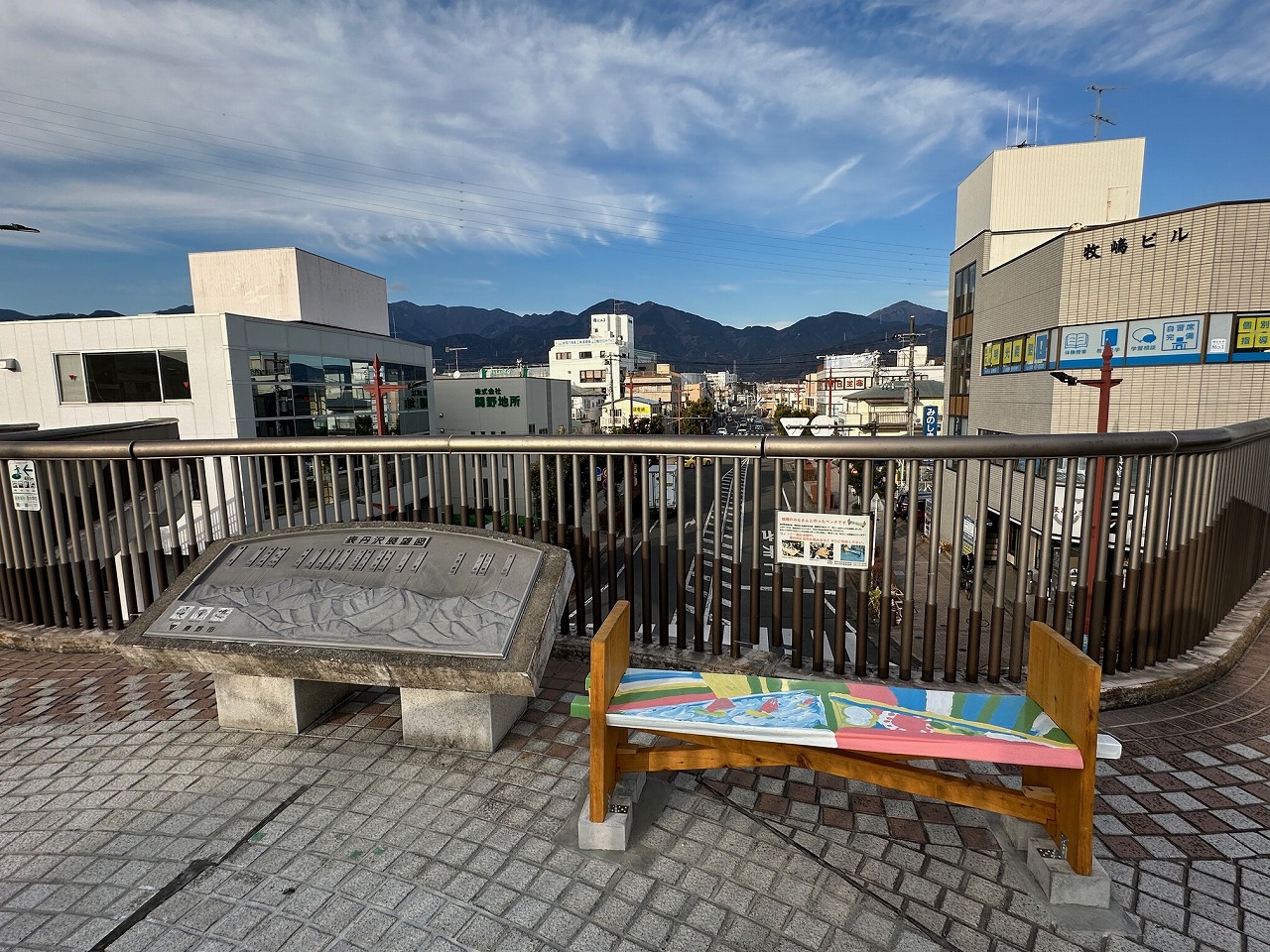 歩道橋の上から街並みと山々を望む景色で、手前にはカラフルなベンチと周辺の立体地図が設置されている写真