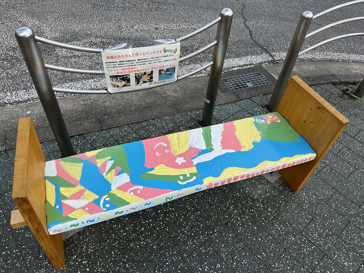 カラフルにペイントされた木製のベンチが歩道に設置され、背面には地域住民が製作に関わった旨の説明文が掲示された写真
