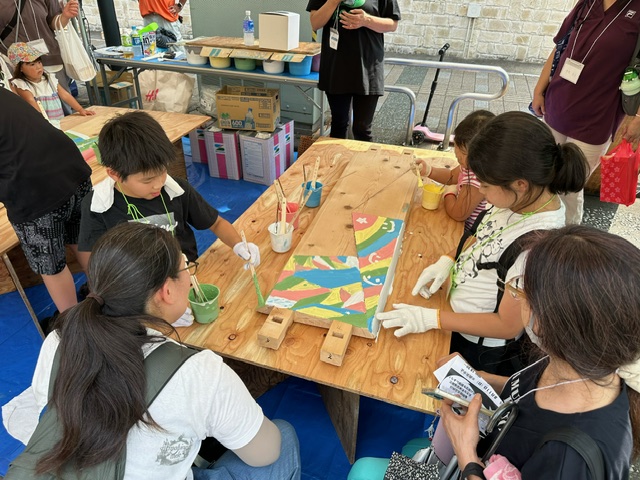 子どもたちと大人が一緒に木製ベンチの座面にカラフルな絵を描いているワークショップの写真