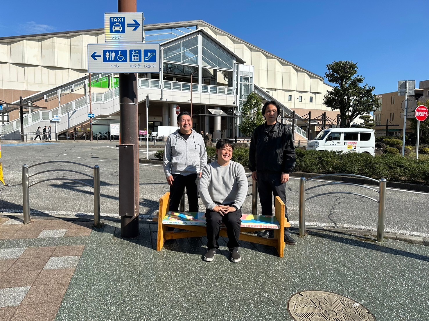 駅前のロータリーで作成したベンチの周囲に2人の人物が立ち、1人がベンチに座っている写真