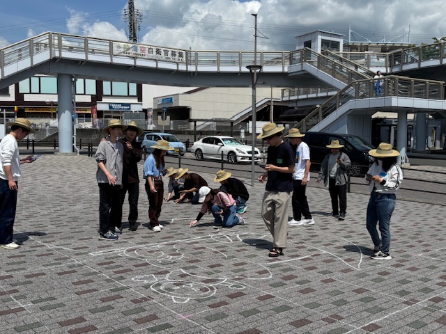 駅前広場らしき舗装された地面で、麦わら帽子を被った十数名の若者たちが集まり、チョークを使って地面に大きな絵を描くアート活動に取り組んでおり、背景には自動車と歩道橋の階段が見えるイベントの様子の写真
