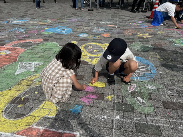 キャップを被った男の子とワンピースの女の子の二人の子供がしゃがみ込み、周囲に既に描かれているカラフルなチョークアートの中で、自分たちの絵を熱心に描いているイベントの様子の写真