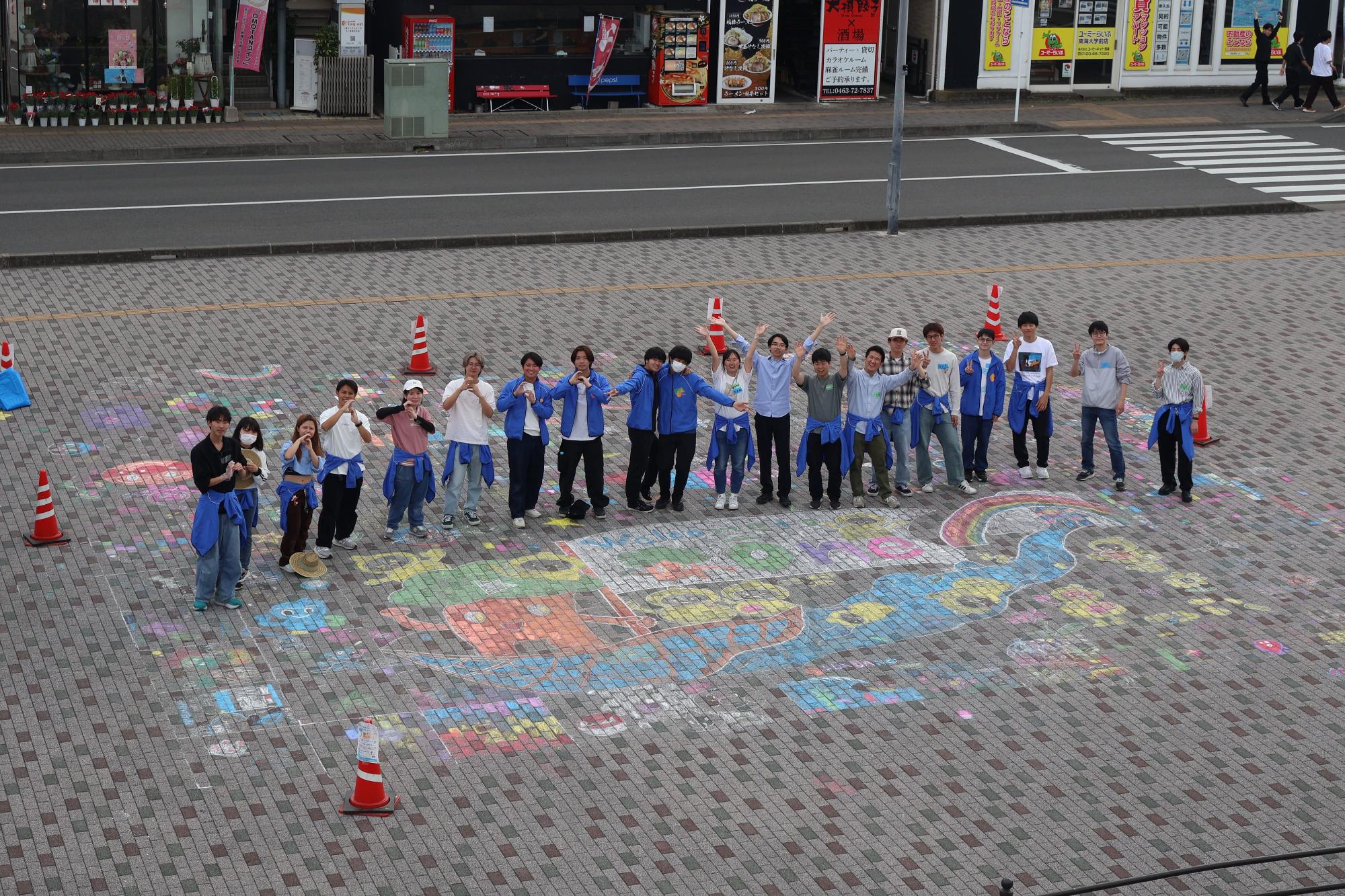 レンガ敷きの屋外広場で、自分たちが描いた巨大でカラフルなチョークアートの周りに、青いパーカーやカジュアルな服装の若者たちが円弧状に並び、手を振ったりピースサインをしたりしてカメラに向かってポーズを取っている集合写真