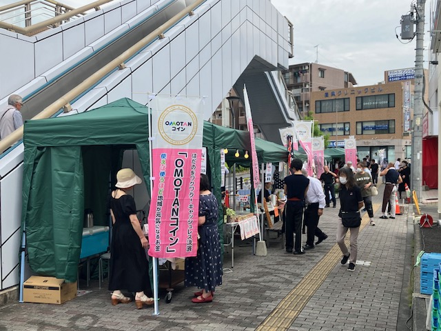 駅の高架下の歩道で開催されている屋外イベントの様子を写した写真で、左側の緑色のテント前には「OMOTANコイン」と書かれたのぼり旗が立てられ、数軒の出店が並び、マスクを着用した人々が買い物を楽しんでいる写真