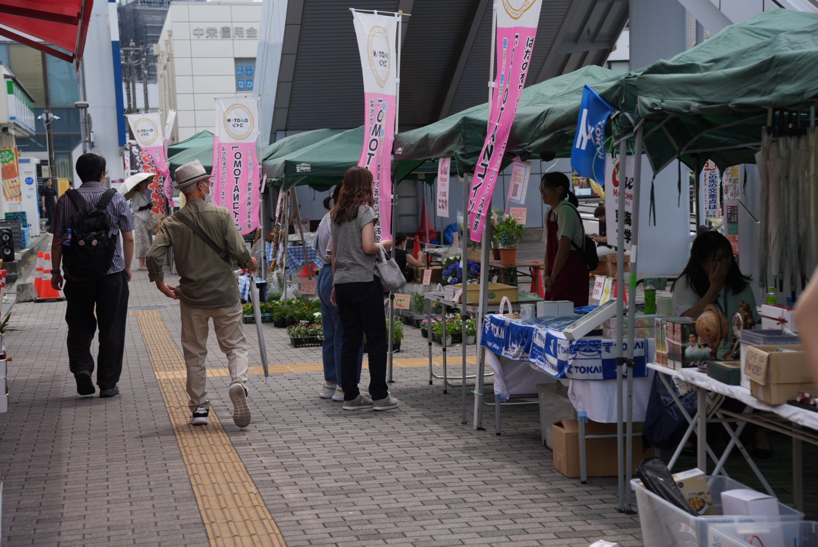 緑色のテントとピンク色ののぼり旗が連なるブースの横を、麦わら帽子を被った年配の男性ら複数の人々が歩いており、ブースでは雑貨や植木などが売られている様子の写真