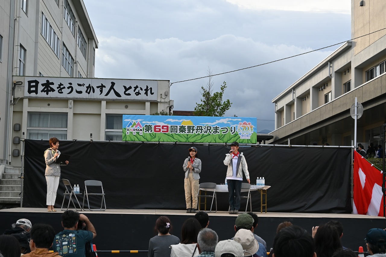 「第69回秦野丹沢まつり」と書かれた看板と「日本をうごかす人となれ」と書かれた横断幕の下に設置された特設ステージの上で、中央の二人がマイクを持って話し、左端の女性が資料を読み上げている写真