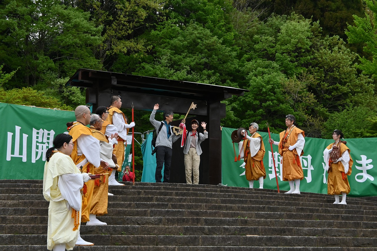 石段の上にある木製の門の前で、黄色や橙色の伝統的な衣装を着た人々が向かい合うように並び、中央で参加者二名が、手を振っている祭事の様子の写真
