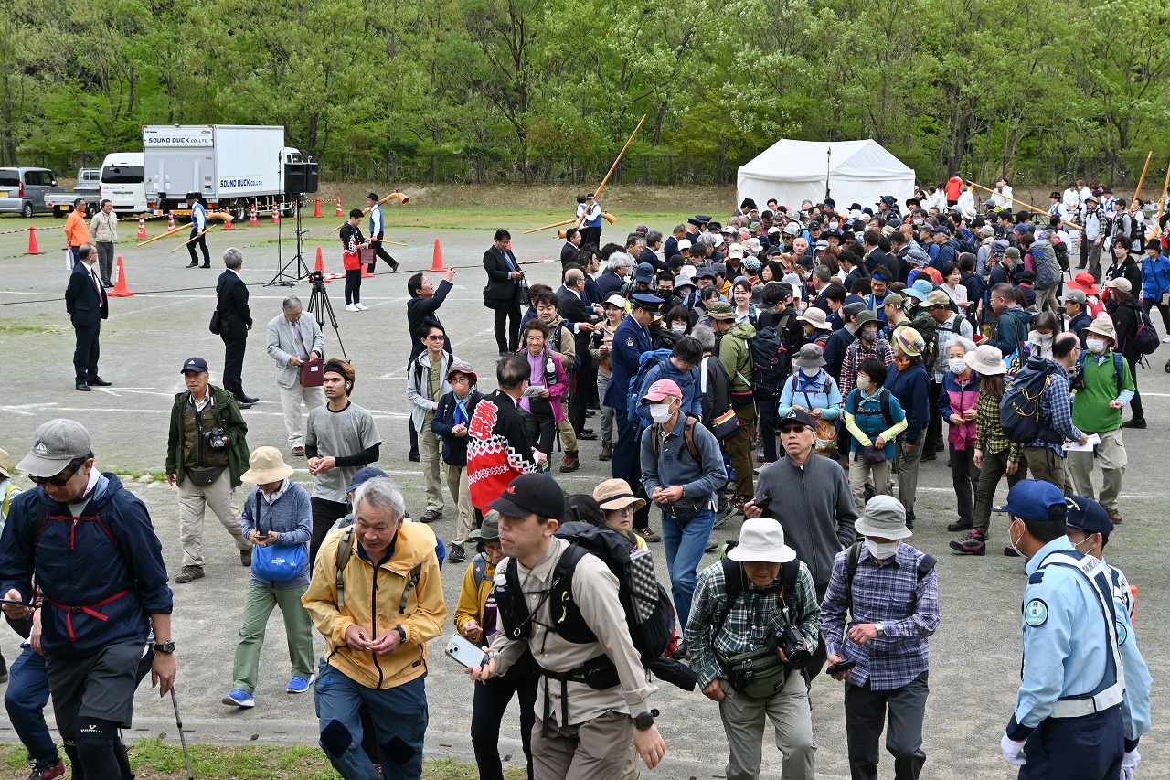 会場に帽子や登山用のアウトドアウェアを着用した大勢の参加者たちが集まり、後方には白いテントや業務用車両、運営スタッフがいるイベントの様子の写真