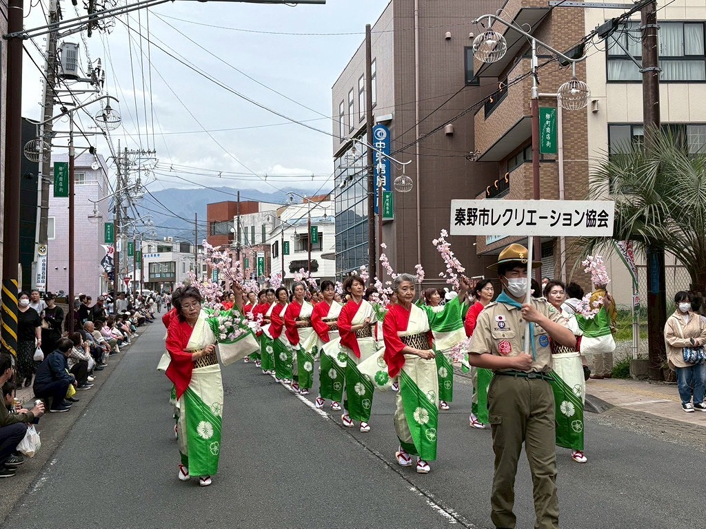 街路で赤と白の着物に緑の裾模様を合わせた衣装を着た女性たちが、桜の飾りを持って踊り、手前には制服を着たボーイスカウトが先頭に立ち行進しているパレードの写真