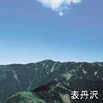 青く澄んだ空に白い雲が一つだけ浮かび、その下には濃い緑と岩肌が露出した高くて険しい山々の稜線が広がっている表丹沢の写真