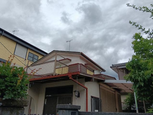 青空を背景に緑に囲まれた高台の白い二階建て住宅が、格子状の手すりとブロック塀の上に立っている写真