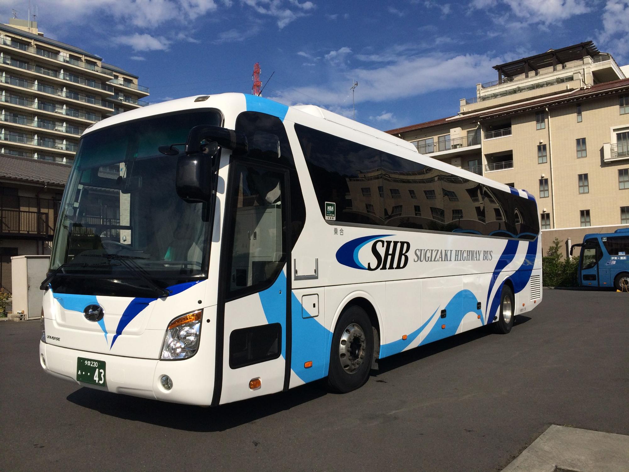 青空の下、住宅やビルが立ち並ぶ駐車場に停車している、白地に青いラインが入り「SHB SUGIZAKI HIGHWAY BUS」と書かれた大型観光バスの写真