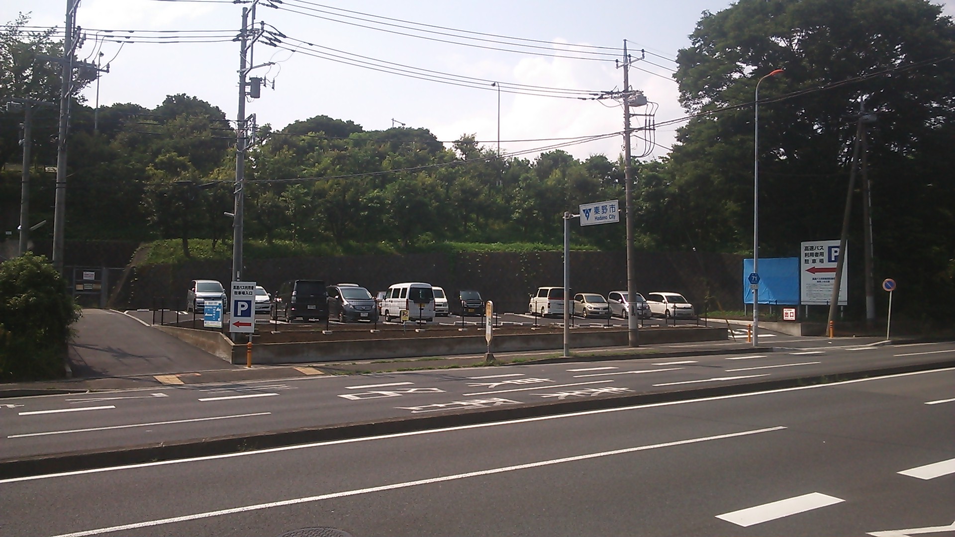 緑豊かな木々を背景に、広い道路の向こう側に複数の白い車両が駐車された駐車場と、青い案内看板や「秦野市」の標識が立つ駐車場付近の写真