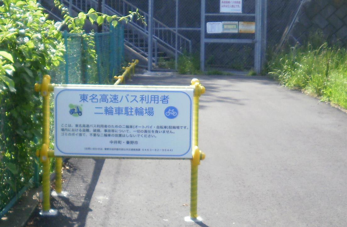 緑の植物に囲まれた通路の入口に、黄色い支柱で支えられた「東名高速バス利用者二輪車駐輪場」と書かれた青い案内看板が設置されている写真