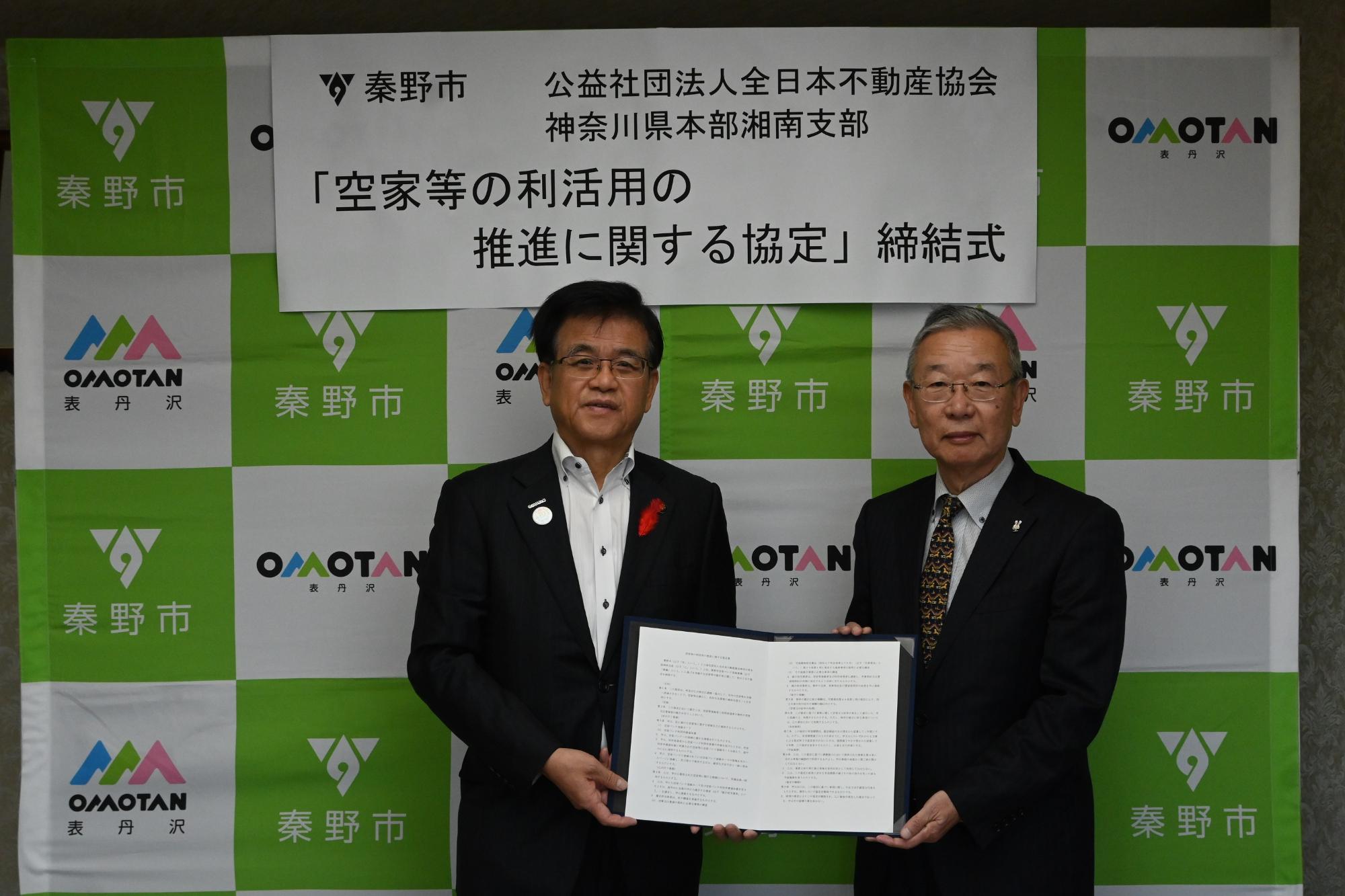 緑と白の秦野市ロゴ入り背景幕の前で「空家等の利活用の推進に関する協定締結式」と掲げられ、スーツ姿の秦野市長と全日本不動産協会神奈川県本部湘南支部代表が協定書を掲げる公式調印式の記念写真