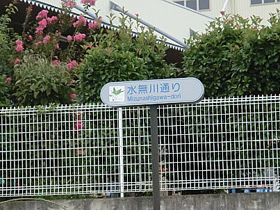白の金網フェンスを背景に、灰色のポールに設置された楕円形の案内標識に「水無川通り」の文字が記された写真