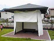 中にベンチが設置されてた、黒い屋根の四方を白い防災シート使用時に覆われた、あずまやの写真