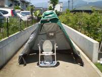 コンクリートの壁に挟まれた通路上のマンホールの上に簡易式の緑色のテントが張られ、中に折りたたみ式の洋式トイレが設置されて災害用緊急トイレの写真