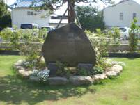 芝生に囲まれた公園の広場にある小さな石が配置された小さな花壇の中に、一本の木と文字が刻まれている大きな石碑が設置された由来板の写真