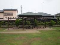 芝生の上に、複数のベンチが配置された休憩所があり、上部にはツタを這わせるための木製の格子が設置されているパーゴラシェルターの写真