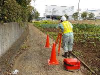 畑の脇の通路で、作業服を着てヘルメットを着用した男性が、カラーコーンを置き、測量をしている様子の写真
