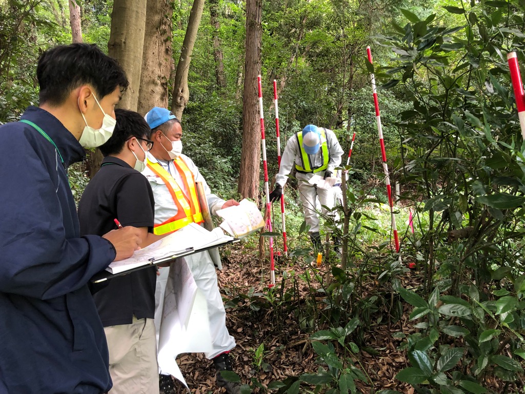 林の中に赤と白の配色のポールを複数設置し、数名の男性が測量を行っている様子の写真