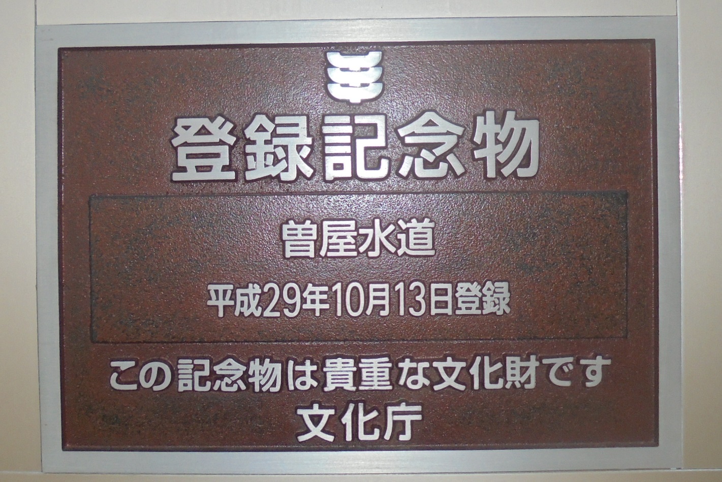 重厚な赤茶色の金属板に、上から「登録記念物」、中央に「曽屋水道」、その下に「平成29年10月13日登録」、最下部に「この記念物は貴重な文化財です」「文化庁」と銀色の文字で刻まれた銘板の写真
