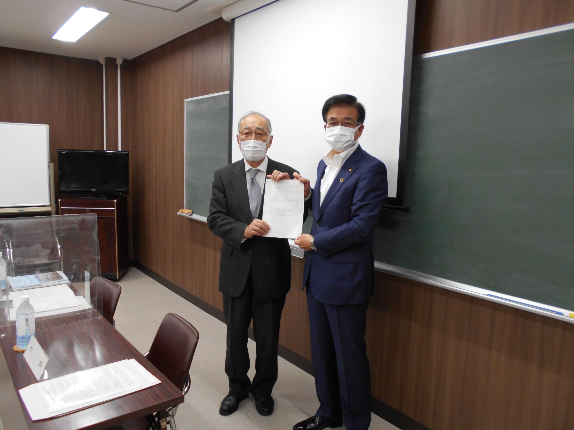 役員室または会議室のような場所で、スーツ姿の二人の男性がマスクを着用し、白い紙の書類を両手で丁寧に持ち、カメラに向かって立っている写真