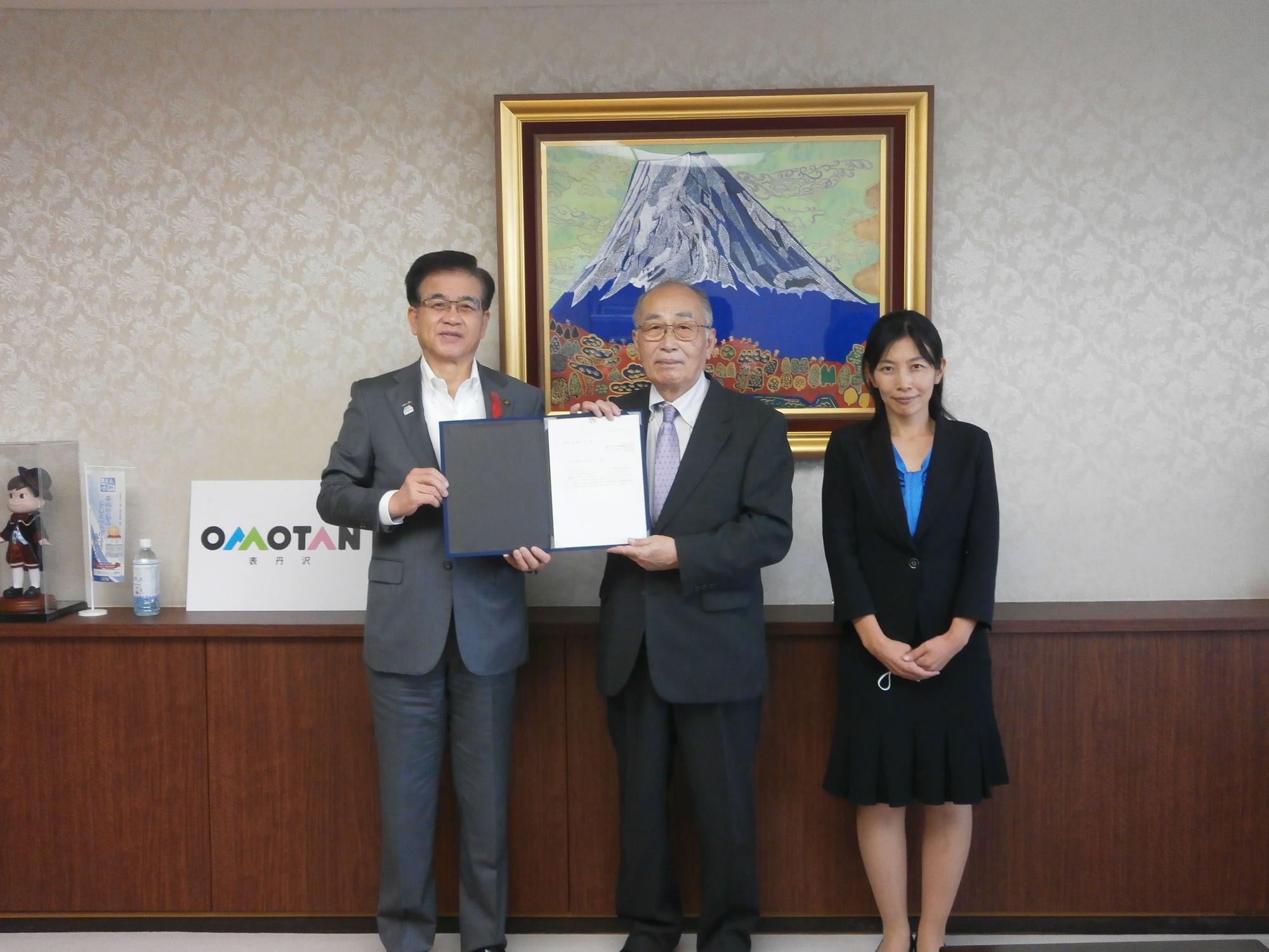 壁紙と木目調のカウンターを背景に、ネクタイを着用した二人の男性と、黒いスーツ姿の女性の合計三人が正面を向き、中央の二人が左右で大きな黒いファイルに挟まれた白い書類を一緒に持っている様子を捉えた写真