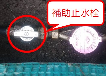水道管の途中に設置された白いT字型のハンドルを持つ補助止水栓を赤い円で囲み、その名称を吹き出しで示しており、右隣には水道メーターの蓋が見える地中の配管部分を接写した写真