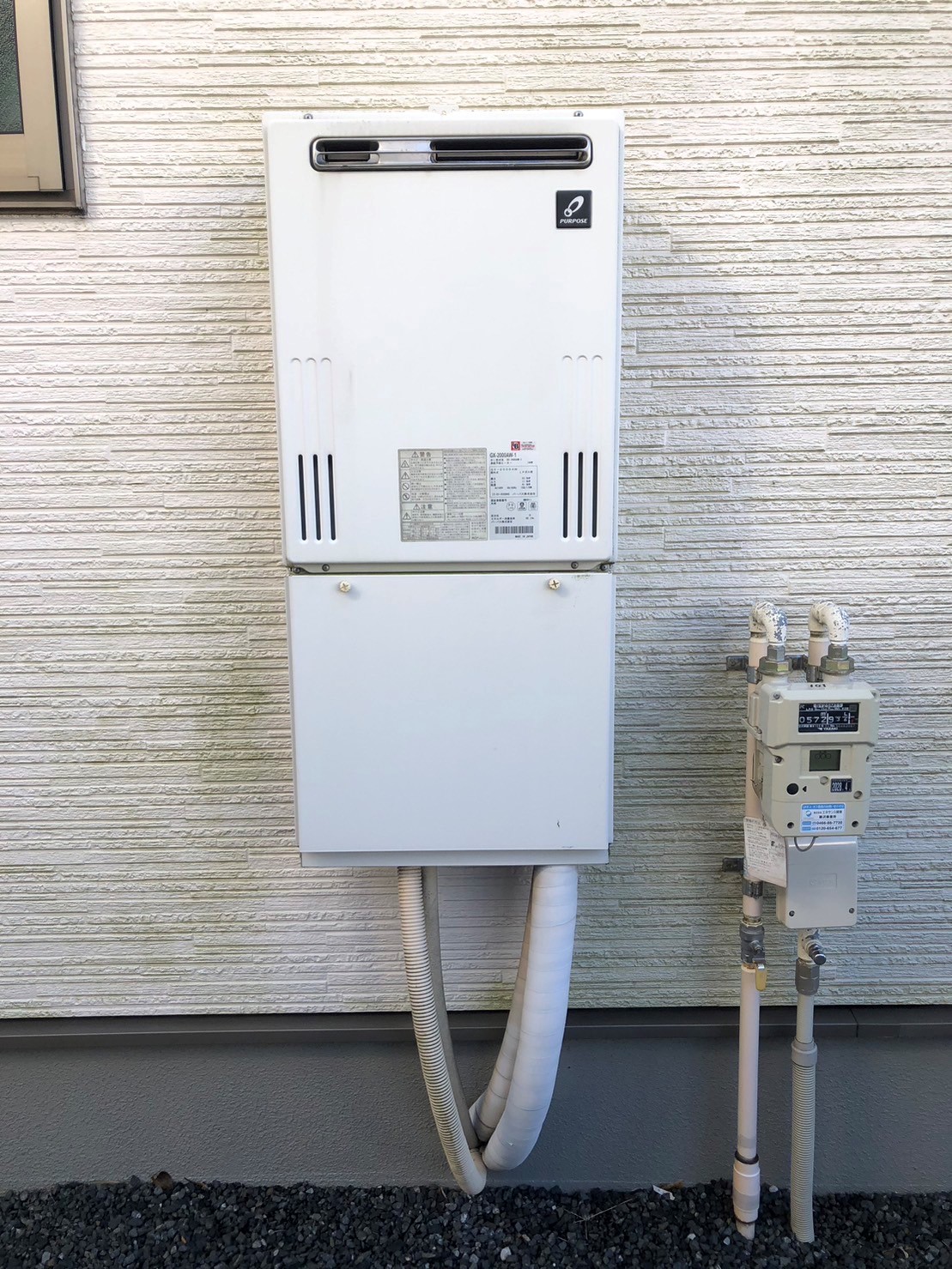 縦長の白い外壁に取り付けられたリンナイ製のガス給湯器の下部から二本の太い白いパイプが出ており、その右隣にはガスメーターや配管が並んでいる屋外機器の設置状況を映した写真