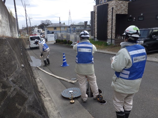 閑静な住宅地の道路で、青いベストとヘルメットを着用した複数の作業員が、開けられたマンホールとそれに接続された白い長いホースの周りで操作訓練を行っている様子の写真