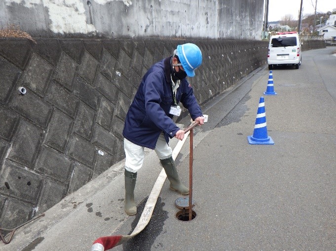 排水路の横の舗装された道路で、青いヘルメットと紺色の作業服を着た男性がマンホールのような穴にレバー状の道具を差し込み、手前に引き寄せている作業の様子の写真
