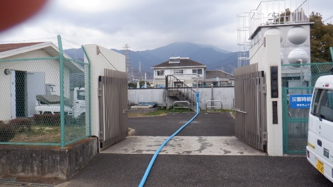 金属製のフェンスと門扉に囲まれた給水場を通る、長い青いホースが敷かれた様子の写真