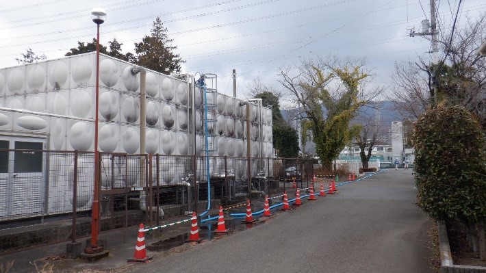 丸い模様が特徴的な大きな白い貯水タンクが金属フェンスの向こうにそびえ立ち、手前の道路沿いには青い給水ホースがオレンジ色のコーンと規制ロープで区切られた落合配水場の全景写真