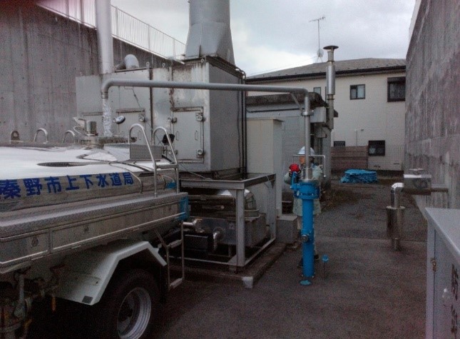 「秦野市上下水道局」と書かれた給水車が、建物の間に設置された銀色の機械設備のそばにある、青い消火栓のような給水口にホースを接続しようとしている様子の写真