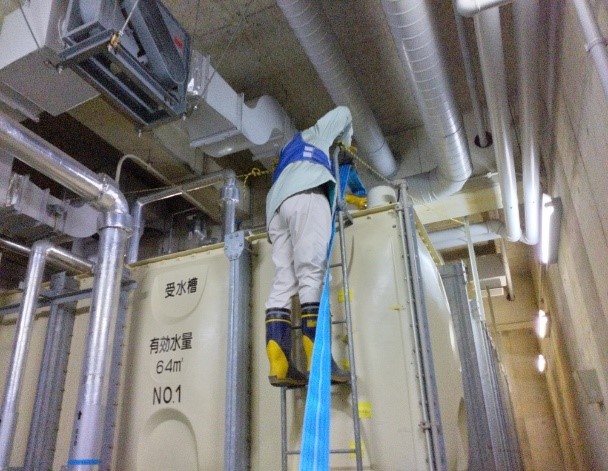 狭い機械室のような空間で、青い安全帯を装着した作業員が白い大きなタンクにかけられた梯子を登り、受水槽へ給水している様子の写真