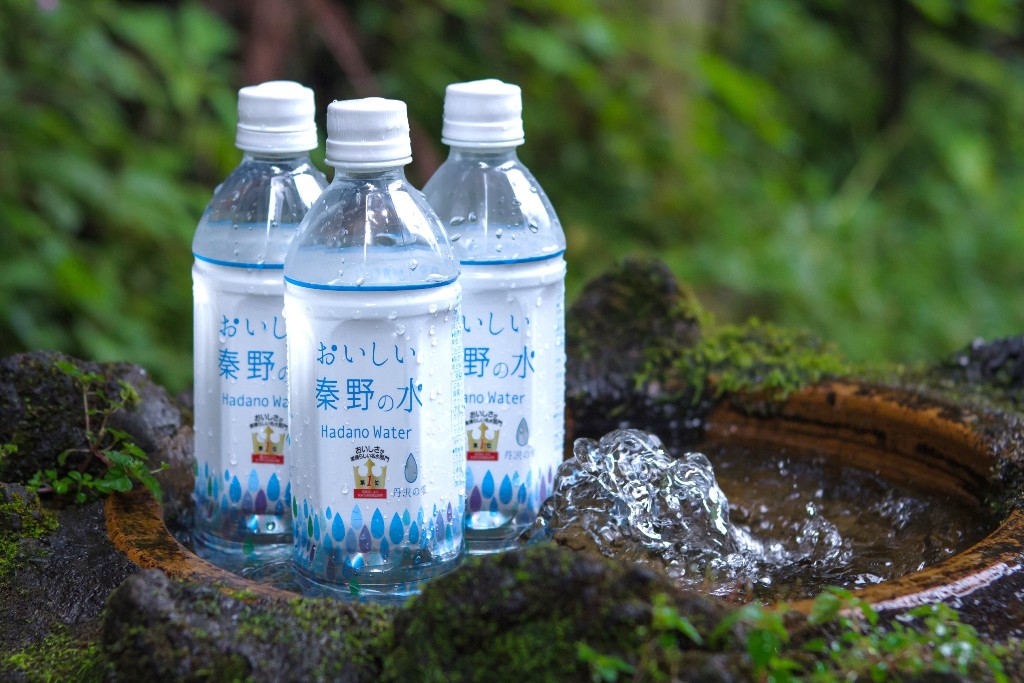 木漏れ日の当たる苔むした石の鉢の周りに、水滴をまとった3本の「おいしい秦野の水 」ペットボトル飲料が置かれ、右下の水面が揺れる静かな自然の中での写真