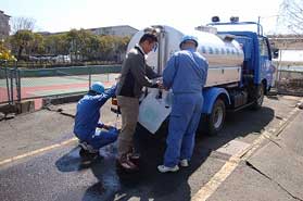 日差しの中、青い作業服の2名の作業員が青い給水車から水をポリタンクへ給水しているのを、ベージュのズボンを履いた男性が手伝っている様子の広場での写真