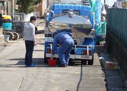 屋外で青い作業服を着た作業員が、後部の銀色のタンクに反射する風景を映す青い給水車から赤いバケツに何かを注ぎ入れる様子を、グレーの服を着た男性が立って見守る作業風景の写真