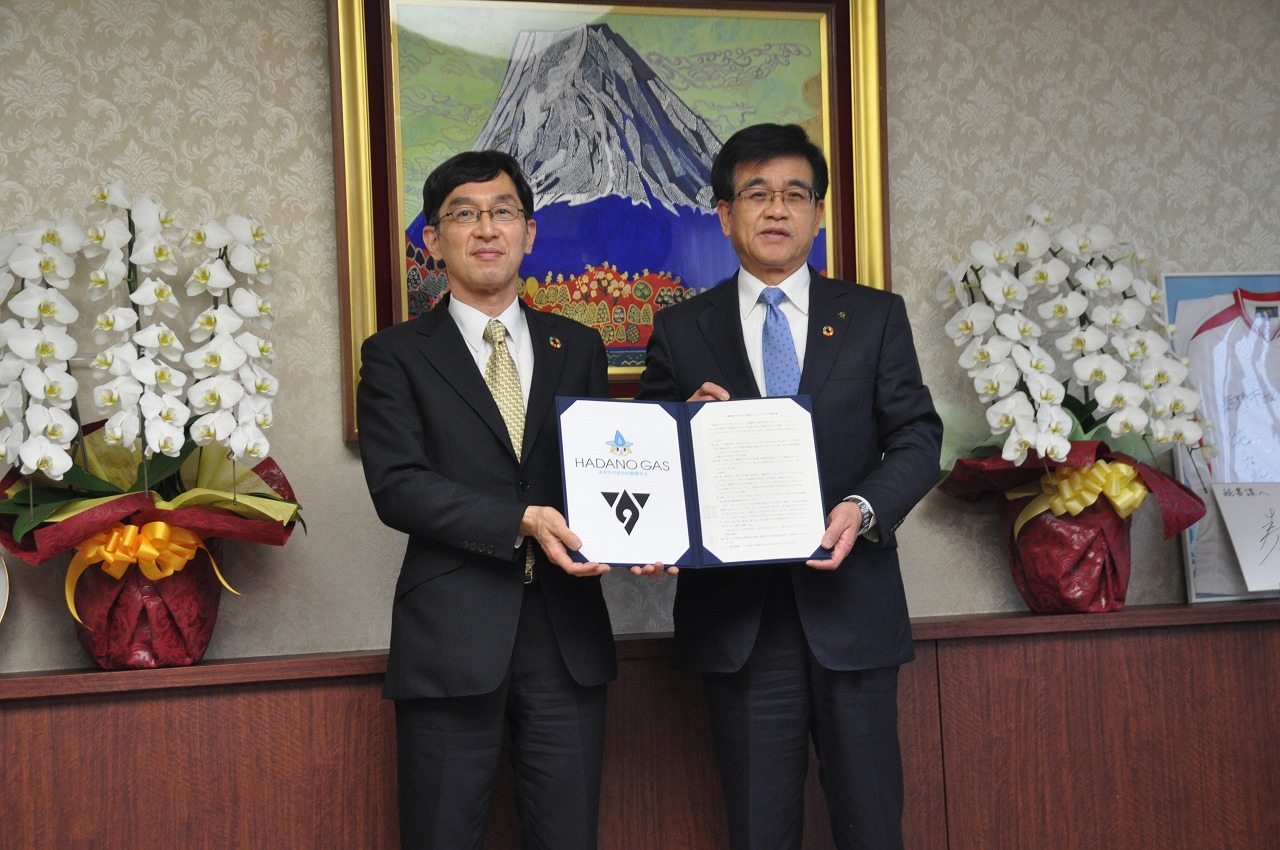 スーツ姿の男性と高橋市長が契約書を一緒に持ち、カメラに見せている様子の写真