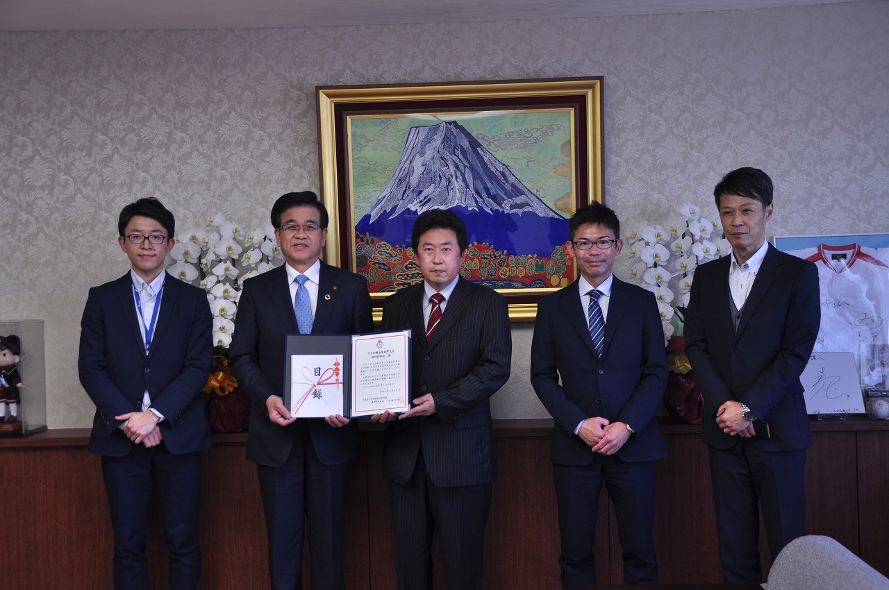 スーツ姿の5人の男性が部屋で正面を向いて立ち、中央の高橋市長と男性が目録と証書ホルダーを一緒に持ち記念撮影を行っている写真