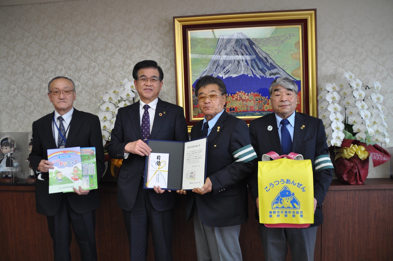 中央の高橋市長と男性が一緒に目録と証書ホルダーを持ち、左側の男性がパンフレットの様な物、右側の男性がランドセルにつけたランドセルカバーの交通安全啓発品をカメラに向け記念撮影を行っている写真