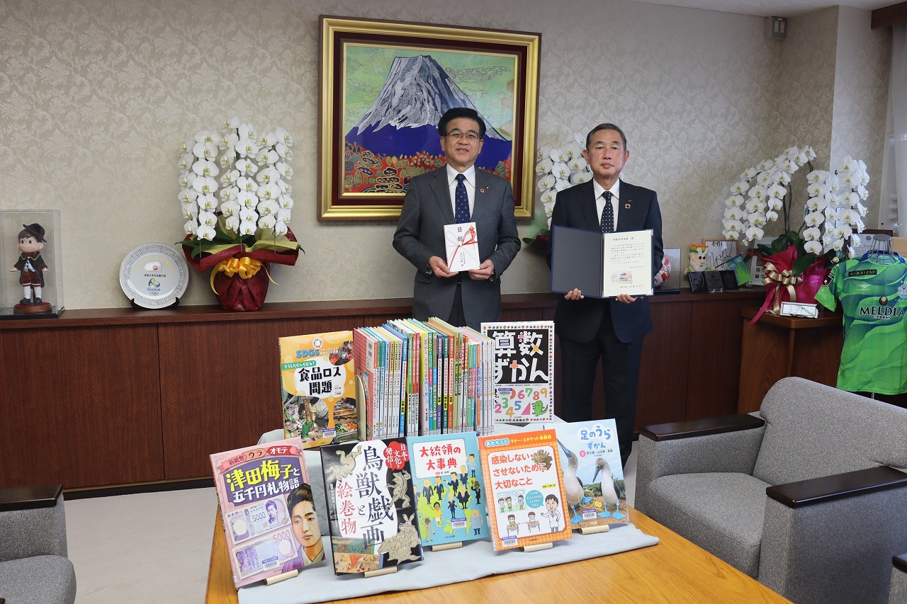 たくさんの絵本や書籍が並べられているテーブルを前に、高橋市長が目録、曽我専務理事が証書ホルダーを持ち記念撮影を行っている写真