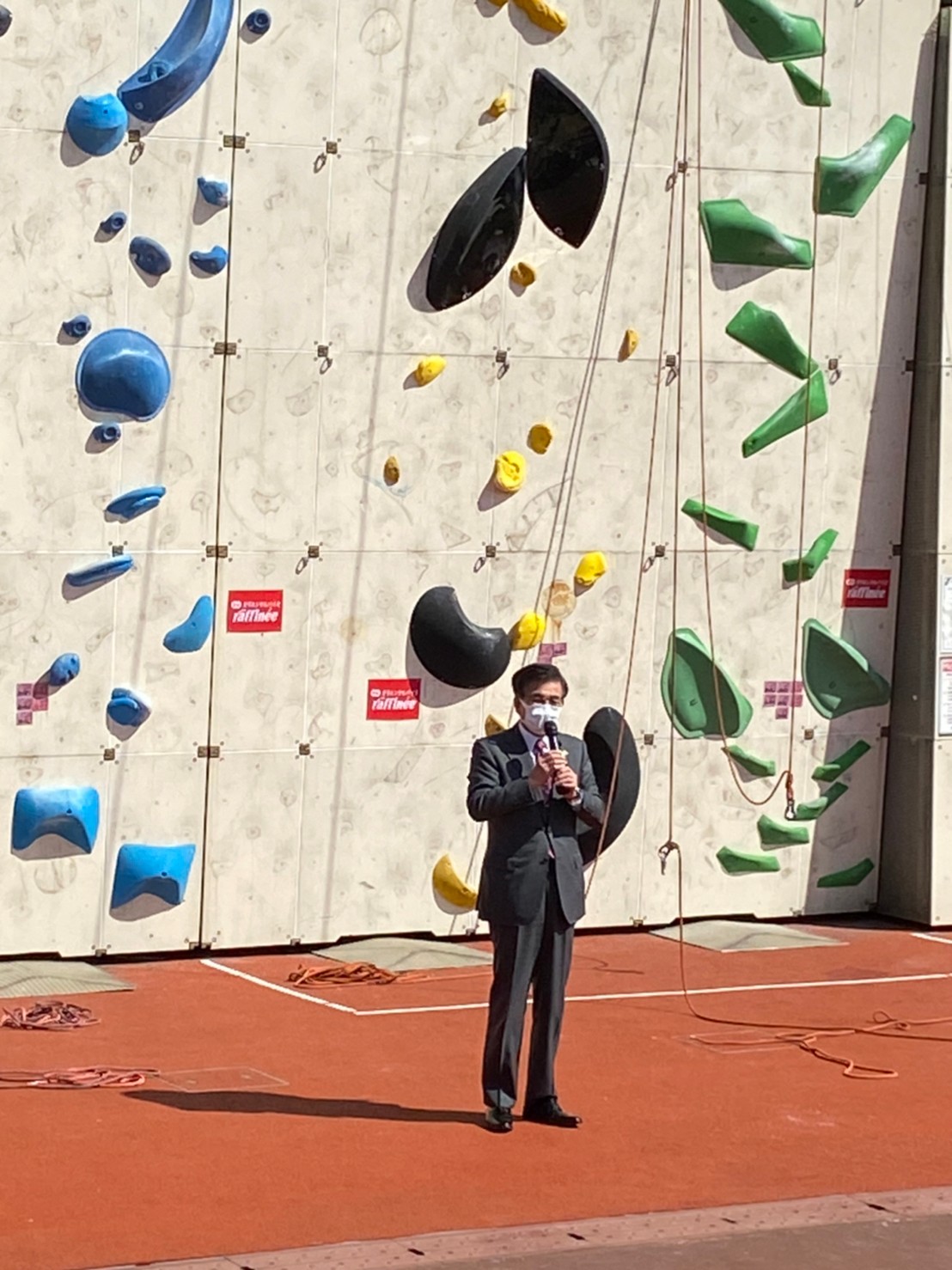 高さのあるボルダリングウォールの前で、高橋市長がマイクを持って、カメラに向かって挨拶をしている様子を捉えた写真