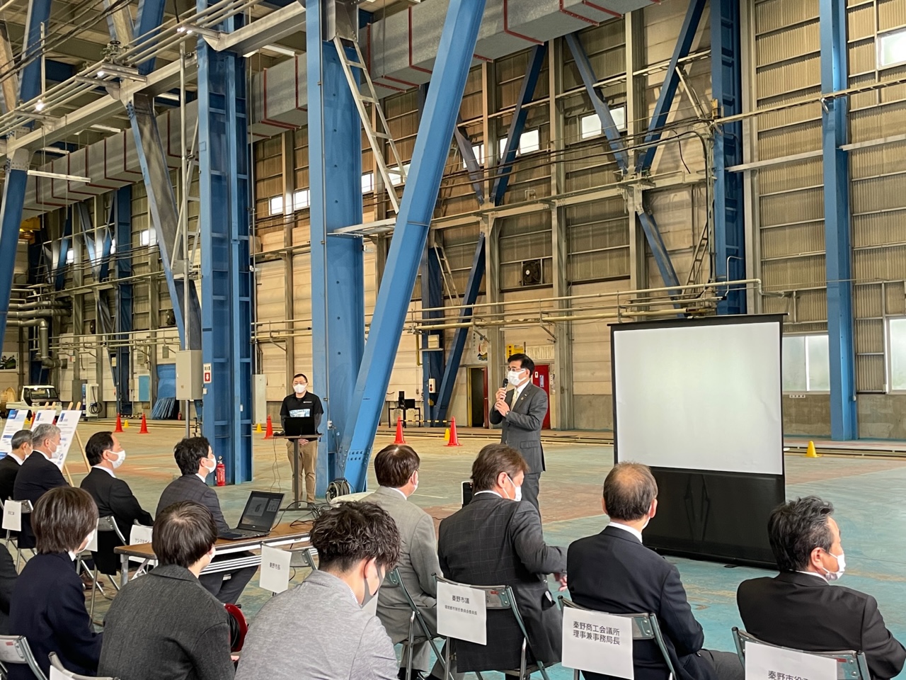 大きな工場の建物内で、スーツ姿の高橋市長がマイクを持ってスピーチをし、複数の人々が椅子に座って話を聞いている写真