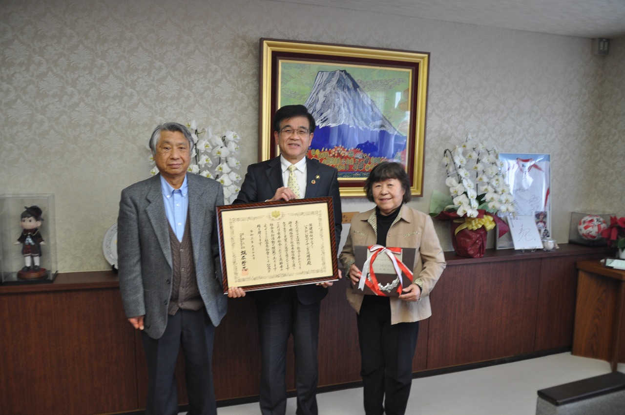 中央の高橋市長と左側の男性が額縁に入った賞状を一緒に持ち、右側の女性が盾を持ち記念撮影を行っている写真