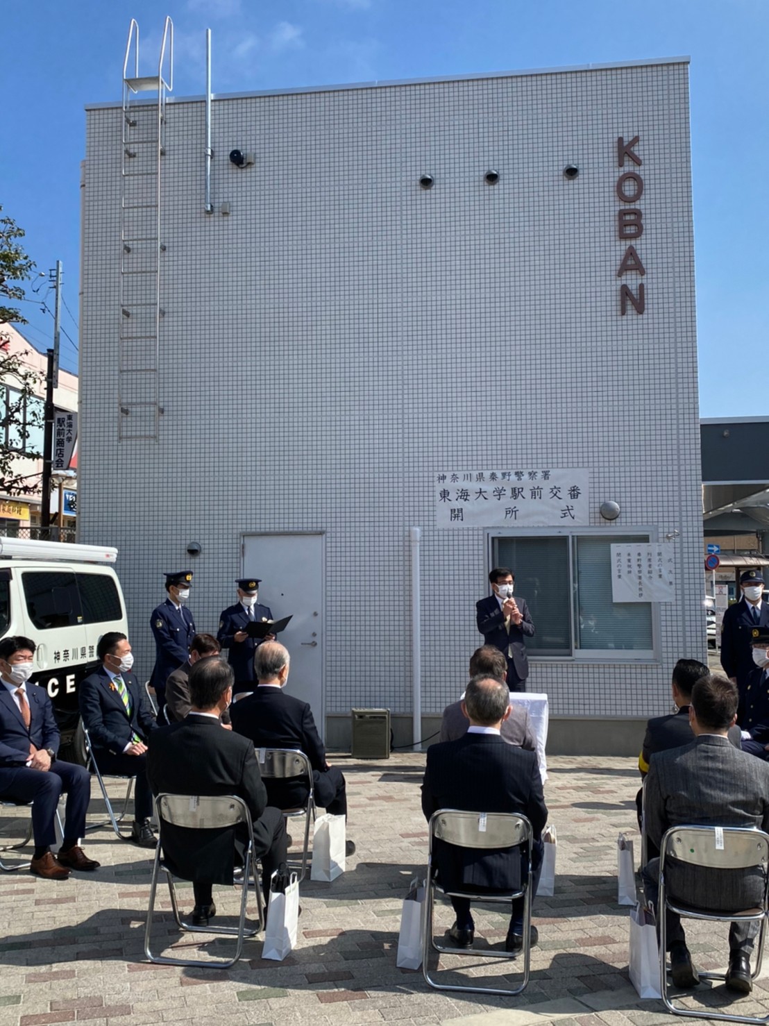 警察官やスーツ姿の男性が椅子に座り、白い壁の交番前でマイクを持つ高橋市長の挨拶を聞く式典の様子の写真