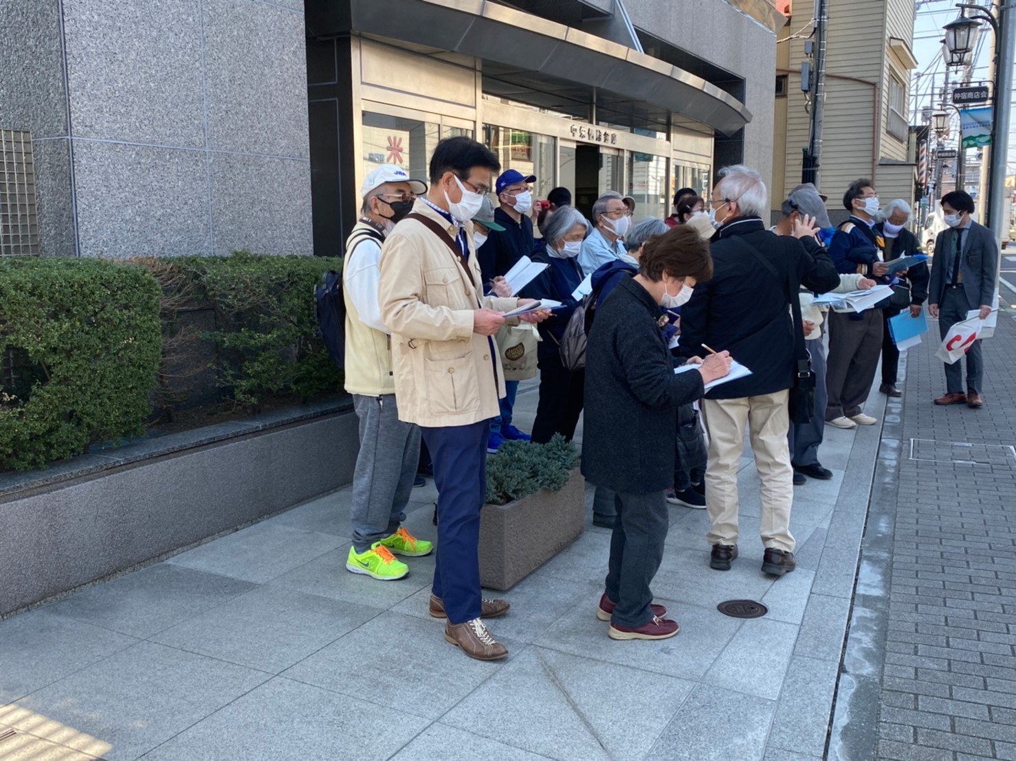 灰色をしたビルの入り口前に参加者たちが集まり、手に持った用紙にメモを熱心にとっている青空レクチャーの様子の写真
